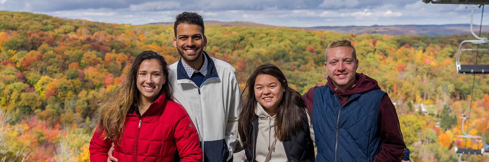 Four friends pose for a picture at the top of the slopes next to a chairlift with fall leaves in the background
