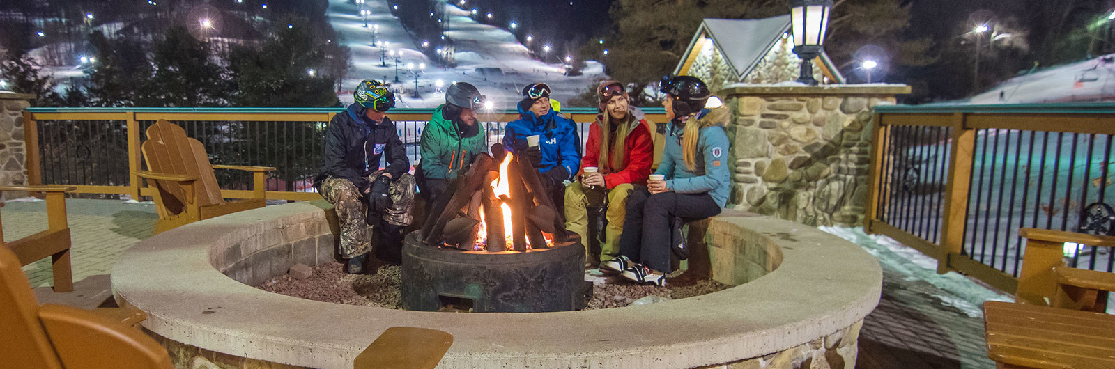Group of skiers and snowboarders sitting around fire pit outside the mountain ski lodge