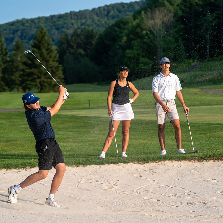 A golfer chipping out of a sand trap with two friends looking on in the background.