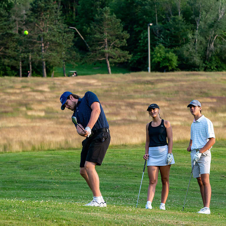 A young man chips a golf ball with two of his friends looking on in the background.