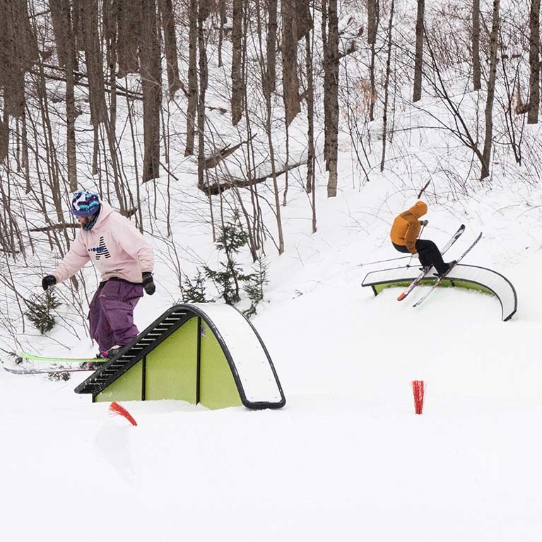 Two skiers sliding on different rails in the terrain park.