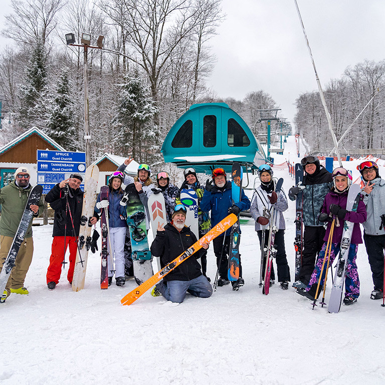 A large group of skiers and snowboarders stands in front of the Spruce chairlift and poses for a picture on a snowy day.